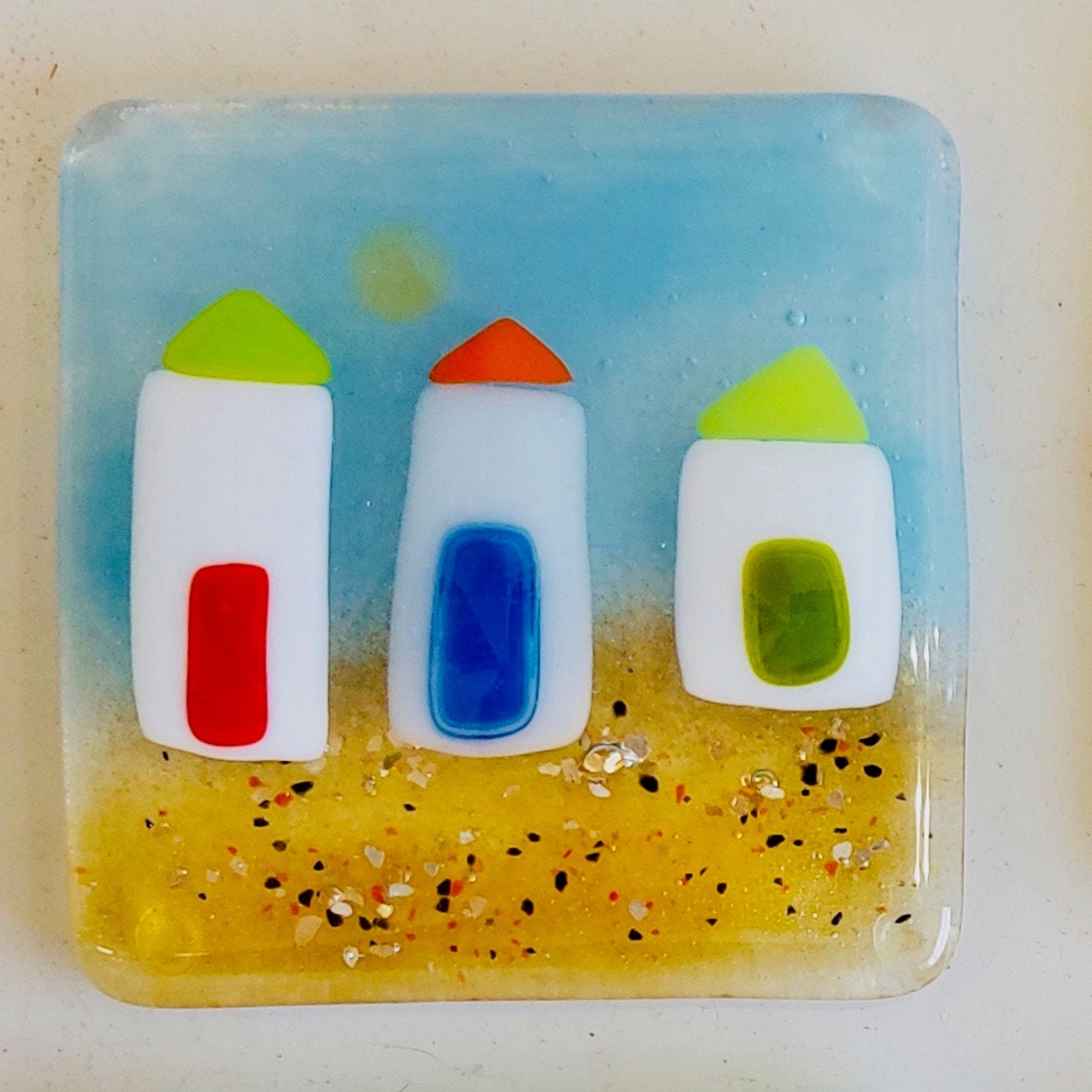 Decorative glass coaster with beach huts on a sandy beach with the sun shining.