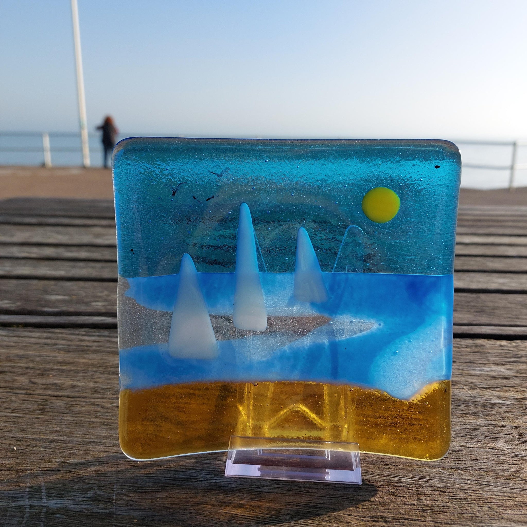Glass artwork depicting a beach scene with a sun and sailboats on a wooden surface.